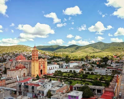 Een luchtfoto van de kleurrijke kathedraal van Tamazulapam in Oaxaca, Mexico, tegen een achtergrond van glooiende groene heuvels en heldere blauwe lucht.