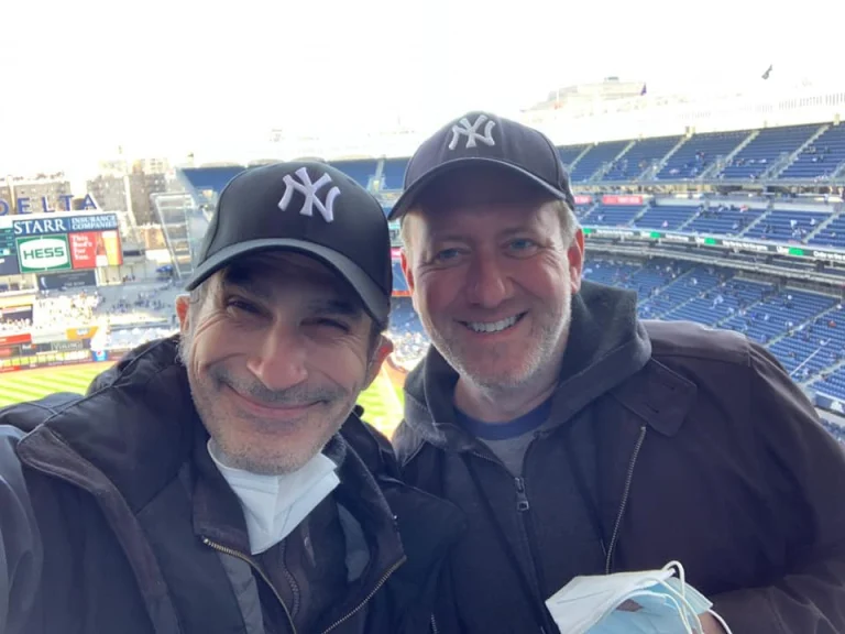 Twee lachende mannen met een blauwe Yankees pet maken een selfie met de tribunes van een honkbalstadion in New York op de achtergrond