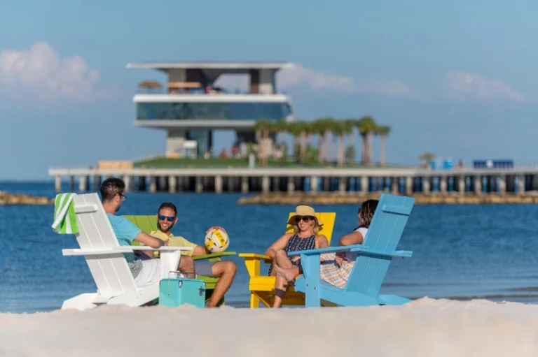 Vier mensen zitten in kleurrijke strandstoelen op het strand met op de achtergrond het moderne gebouw van de St Pete Pier in Florida