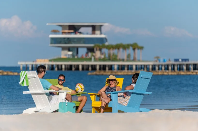 Vier mensen zitten in kleurrijke strandstoelen op het strand met op de achtergrond het moderne gebouw van de St Pete Pier in Florida
