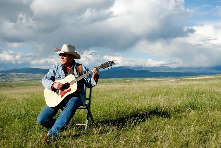 Man met cowboyhoed speelt gitaar zittend op een kruk in een grasveld met bergen op de achtergrond in Montana