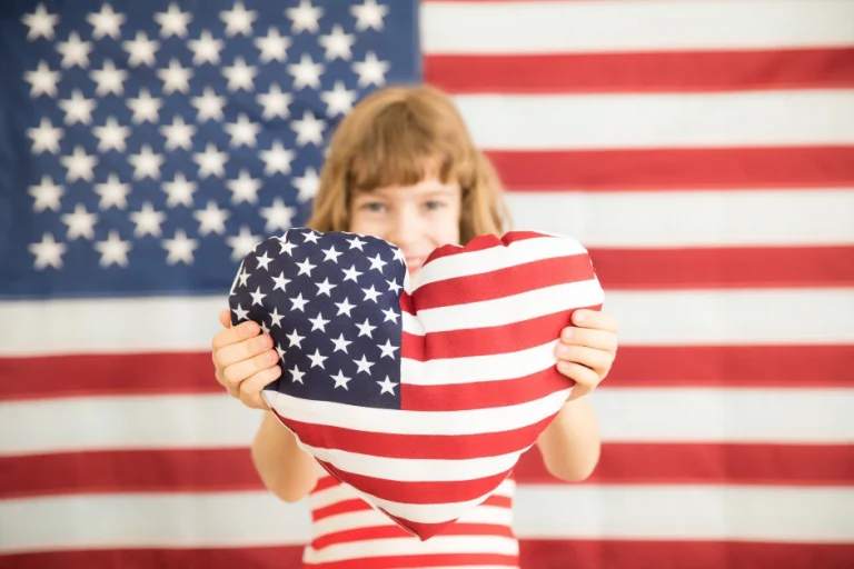 Meisje houdt een hartvormig kussen in de kleuren van de Amerikaanse vlag voor haar gezicht met een grote vlag op de achtergrond