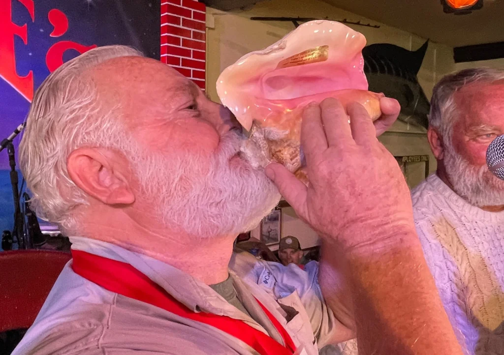 Man verkleed als Ernest Hemingway met witte baard blaast op een grote roze schelphoorn tijdens een festival in Key West