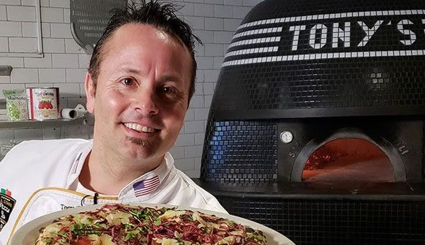 Lachende chef in wit werktenue houdt een pizza omhoog voor een grote zwarte houtoven met het opschrift Tony's in San Francisco