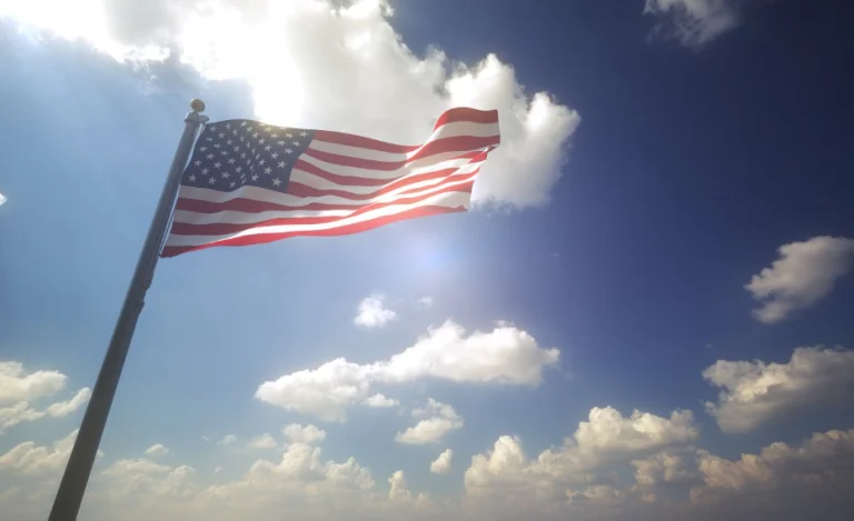 Amerikaanse vlag met sterren en strepen wappert in de wind tegen een felblauwe lucht met stapelwolken en zonlicht