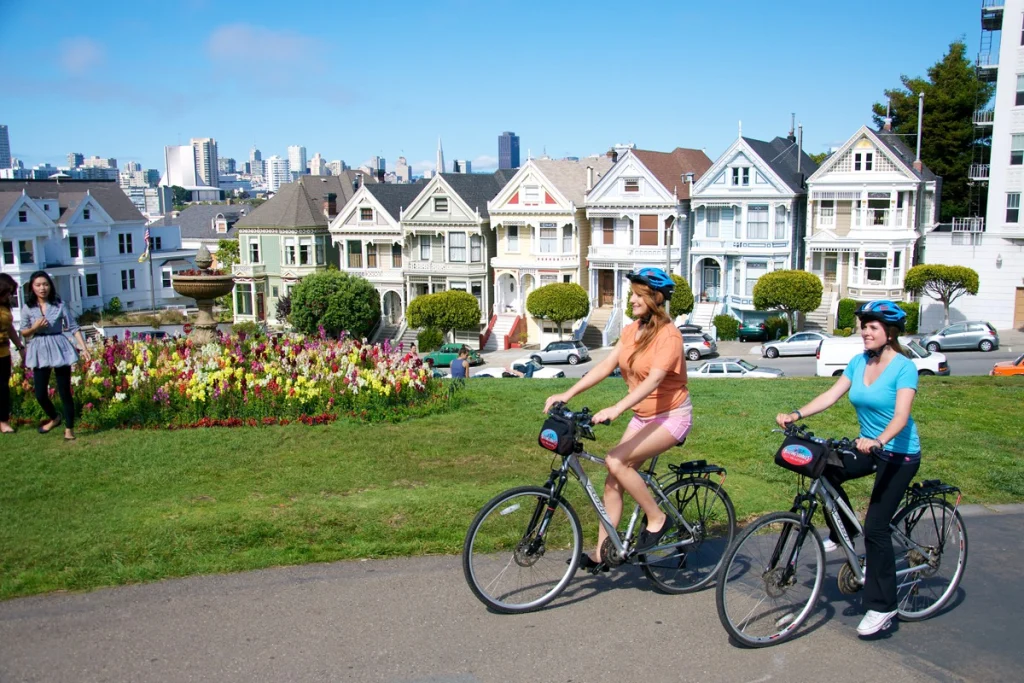 Twee vrouwen met fietshelm fietsen langs een bloemenperk met op de achtergrond de kleurrijke victoriaanse huizen en de skyline van San Francisco