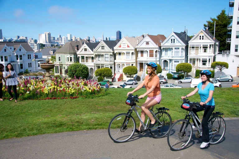 Twee vrouwen met fietshelm fietsen langs een bloemenperk met op de achtergrond de kleurrijke victoriaanse huizen en de skyline van San Francisco