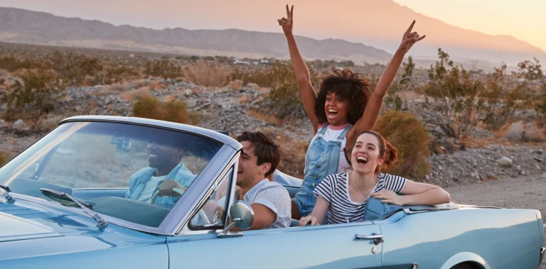 Drie lachende vrienden rijden in een lichtblauwe klassieke cabriolet door een rotsachtig woestijnlandschap bij zonsondergang