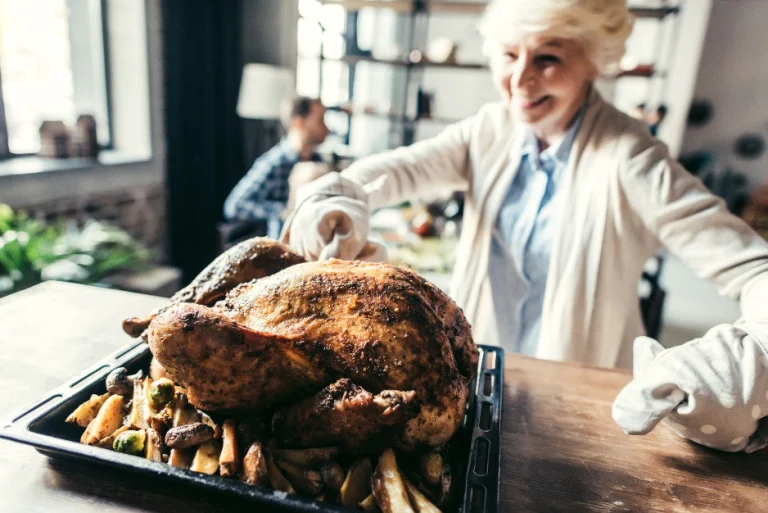 Lachende oudere vrouw met ovenwanten zet een grote gebraden kalkoen met bijgerechten op tafel voor Thanksgiving