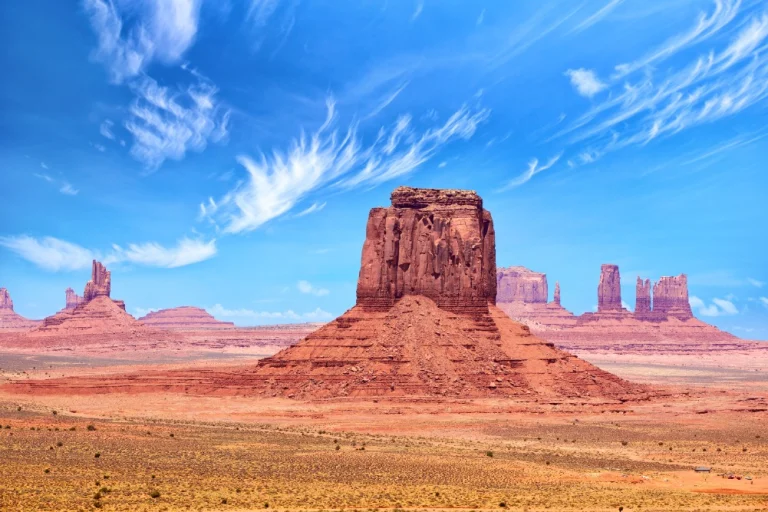 Losstaande rode rotsformatie in Monument Valley met verspreide wolken aan een blauwe lucht en droge grond op de voorgrond