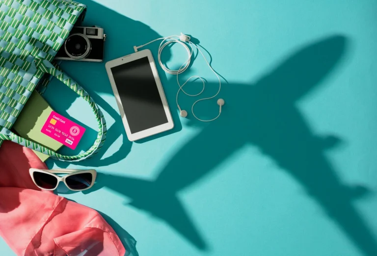 Zonnebril, tablet, camera en tas liggen op een turkooise ondergrond met de schaduw van een opstijgend vliegtuig ernaast
