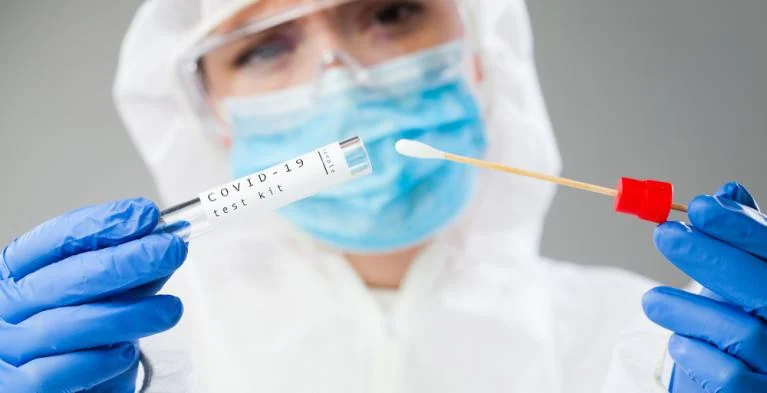 Persoon in beschermende kleding en mondkapje houdt een covid-19 testbuisje en wattenstaafje vast