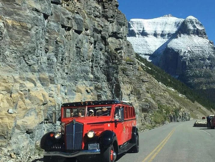 Rode klassieke tourbus rijdt langs een rotswand met besneeuwde bergtoppen op de achtergrond in Glacier National Park