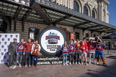 Groep hockeyspelers en teamleden poseert bij een groot rond bord met de tekst Denver 2022 Hockey Capital USA voor het historische Union Station