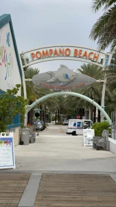 Boogvormige ingang met het opschrift Pompano Beach en een grote vissculptuur boven een promenade met palmbomen