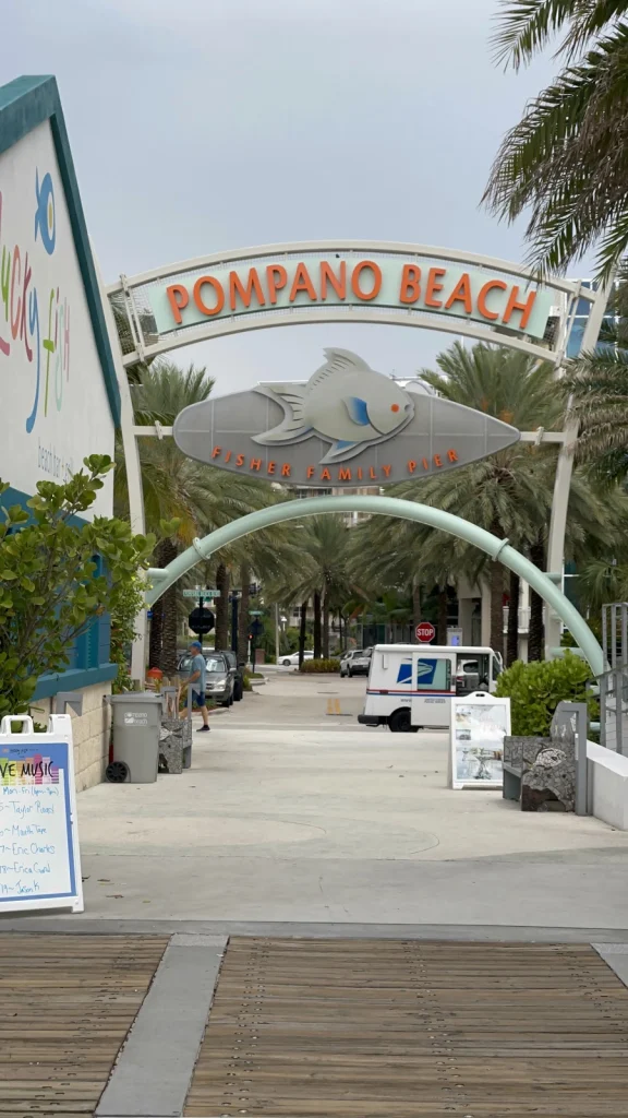 Boogvormige ingang met het opschrift Pompano Beach en een grote vissculptuur boven een promenade met palmbomen