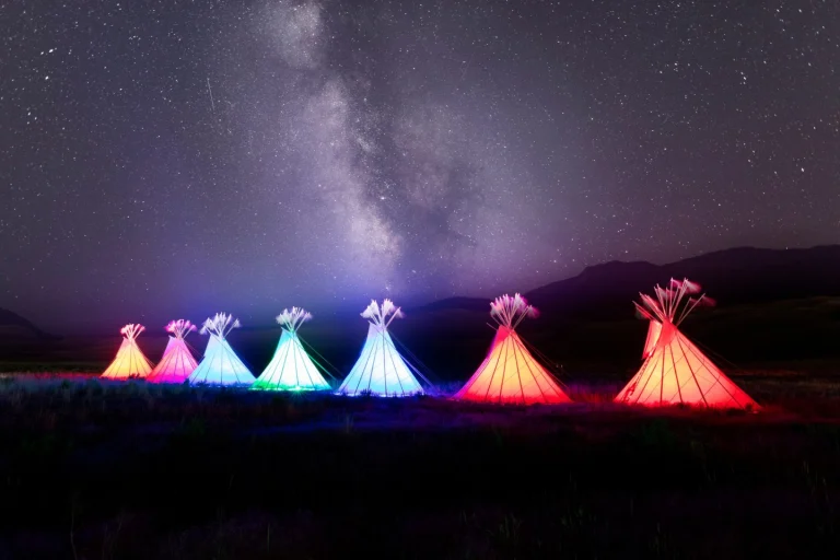 Rij kleurrijk verlichte tipi's op een veld onder een heldere sterrenhemel met de Melkweg zichtbaar in Yellowstone National Park