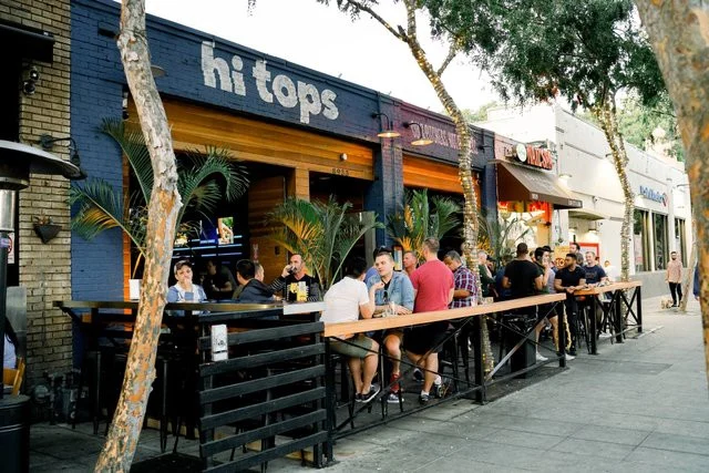 Mensen zitten aan een lange bartafel op het buitenterras van bar Hi Tops tussen palmbomen in West Hollywood
