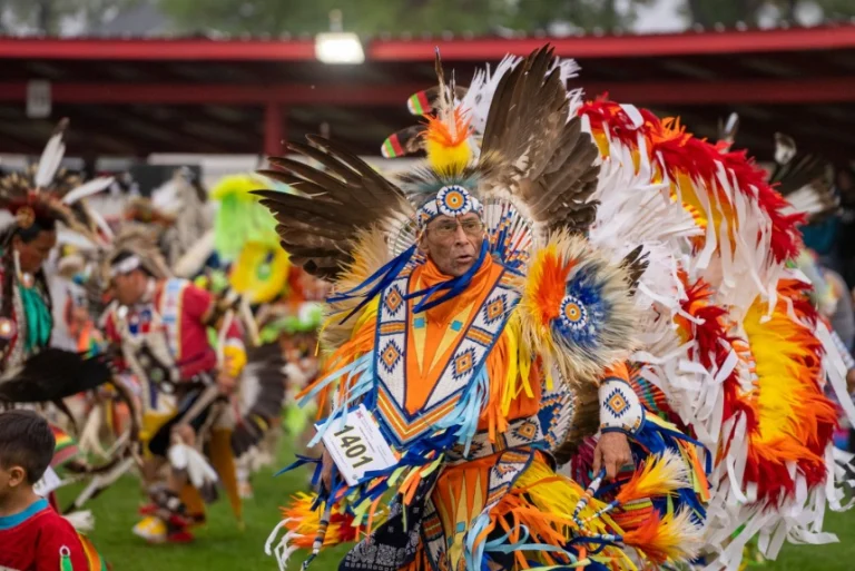 Man draagt een indrukwekkend kostuum met oranje, wit en blauw gekleurde veren en danst tijdens een powwow, met andere dansers wazig op de achtergrond