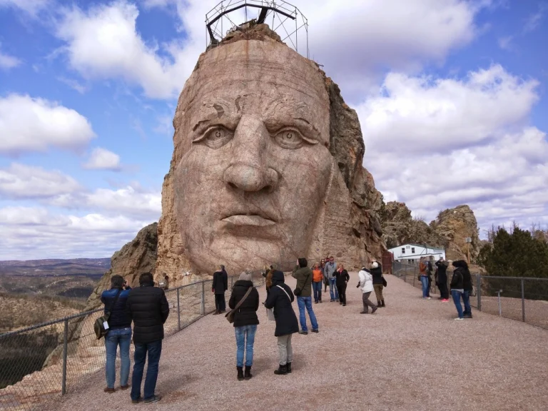 Groep toeristen staat bij een hek en kijkt naar het enorme uit de rots gehouwen gezicht van het Crazy Horse Memorial onder een wolkenlucht