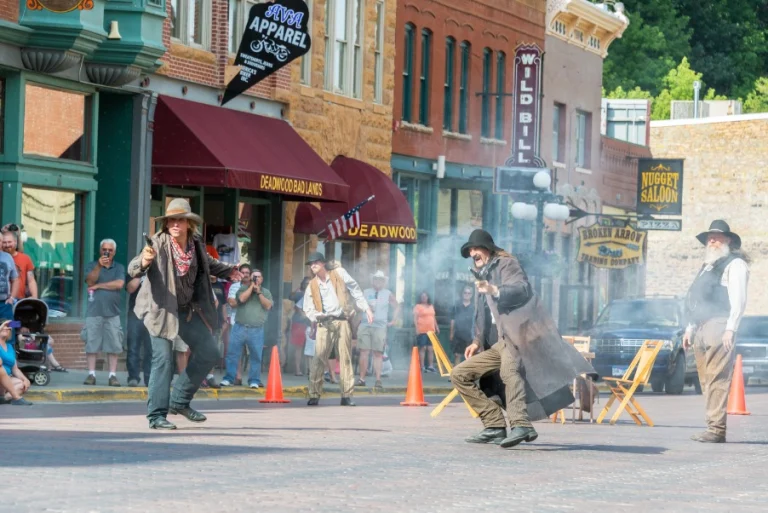 Acteurs in cowboykleding spelen een vuurgevecht na met rooksporen, toekijkende bezoekers en historische gebouwen met saloon uithangborden in Deadwood