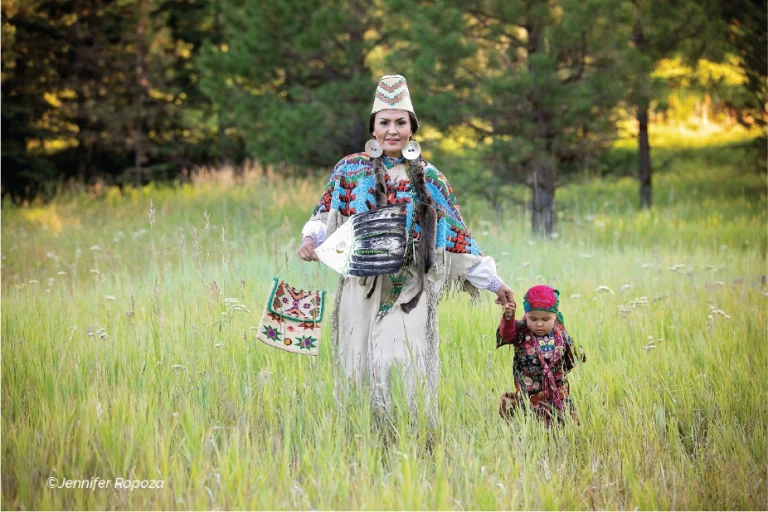 Vrouw in kleurrijke traditionele kleding met kralenwerk loopt hand in hand met een klein kind door hoog gras voor een bosrand