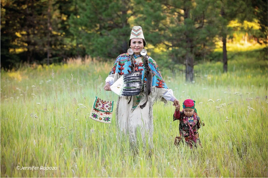 Vrouw in kleurrijke traditionele kleding met kralenwerk loopt hand in hand met een klein kind door hoog gras voor een bosrand