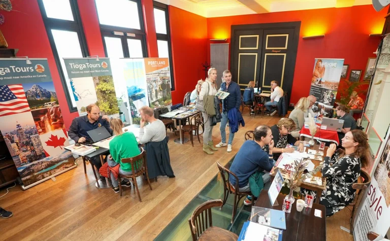 Overzichtsfoto van een reisbeurs met bezoekers aan tafels, reisbanners voor Amerika en Canada en medewerkers die klanten te woord staan in een rode ruimte