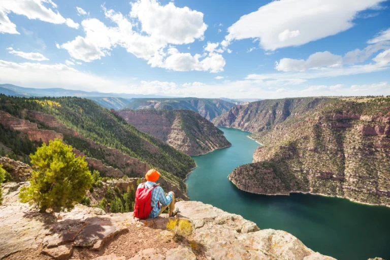 Wandelaar met rode rugzak zit op een rotsrand met uitzicht over het turquoise water van Flaming Gorge Reservoir tussen rode kloofwanden