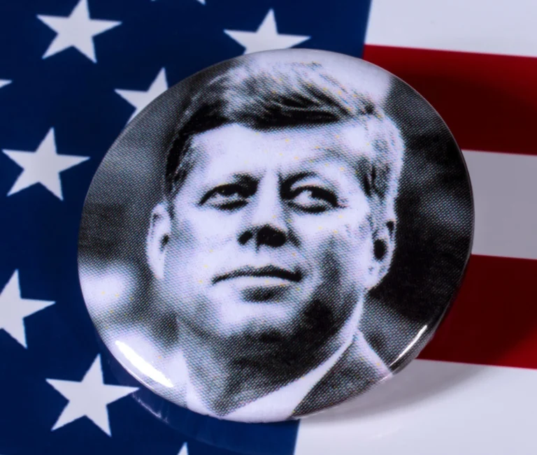 Ronde button met een zwart wit portretfoto van John F. Kennedy liggend op een Amerikaanse vlag met sterren en strepen