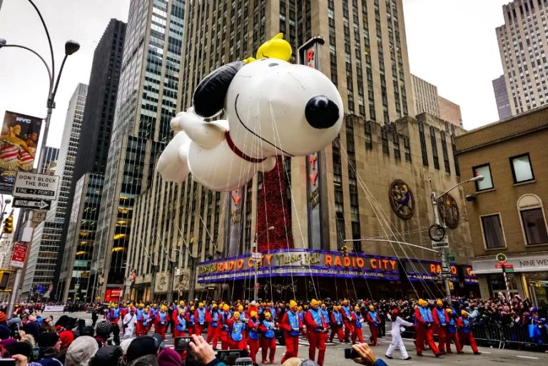 Grote opblaasbare Snoopy ballon met Woodstock erbovenop zweeft door een straat langs wolkenkrabbers, met dansers in kleurrijke pakken en toeschouwers bij Radio City Music Hall