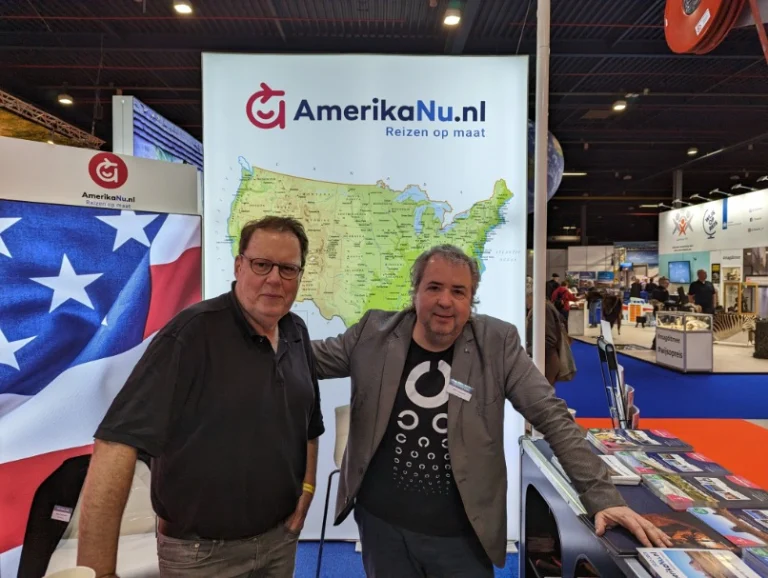 Twee mannen poseren glimlachend bij een standbanner van AmerikaNu.nl met een kaart van de Verenigde Staten en een Amerikaanse vlag op een reisbeurs