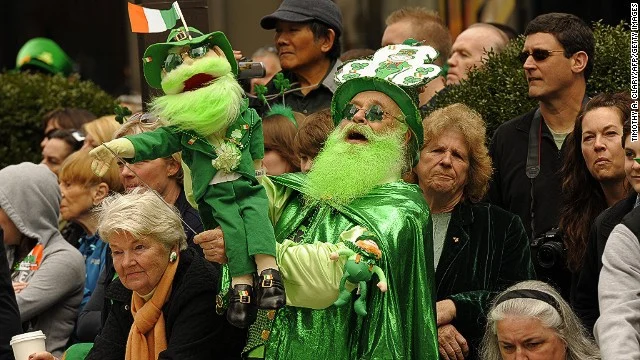 Menigte toeschouwers in groene kleding en met een groene pop kijkt naar de St. Patrick's Day parade, met een Ierse vlag op de achtergrond