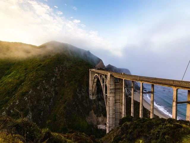 Betonnen boogbrug over een kloof met uitzicht op de kust, met laaghangende mist over de heuvels langs de Californische kustweg
