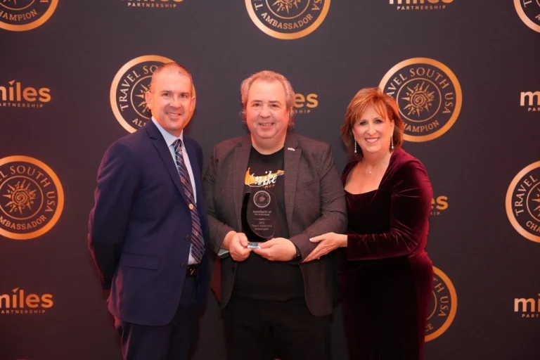 Twee mannen en een vrouw in avondkleding poseren met een glazen trofee voor een backdrop met logo's van Travel South USA en Miles Partnership