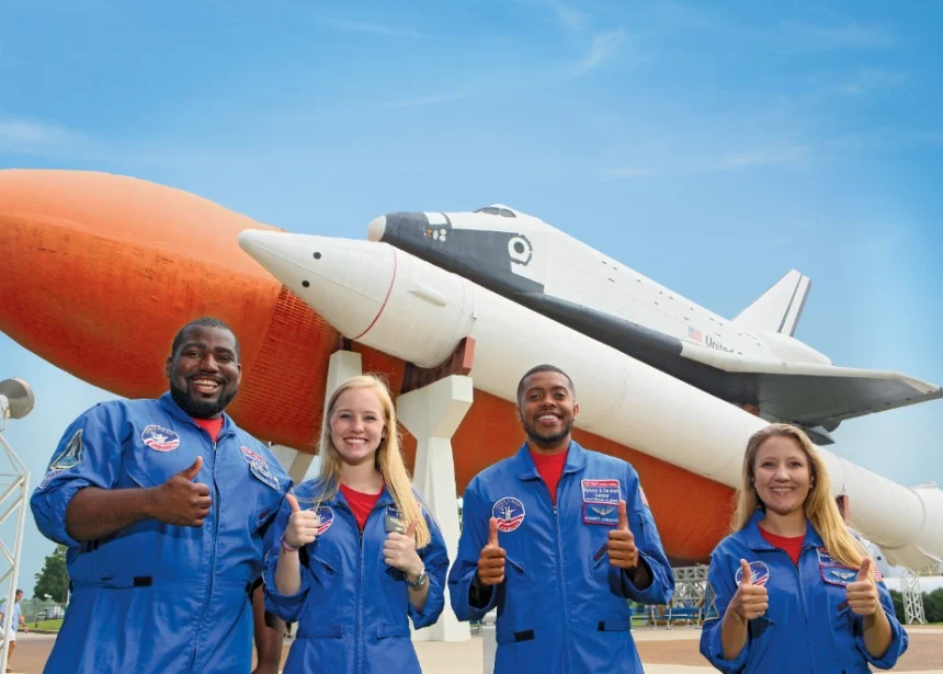 Vier bezoekers in blauwe astronautenpakken steken hun duim op voor een space shuttle en raket in het US Space Rocket Center in Alabama