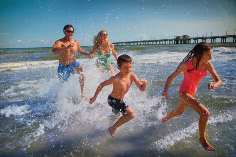 Vader, moeder, zoon en dochter rennen spetterend door de golven op een zonnig strand met een lange pier op de achtergrond in Alabama