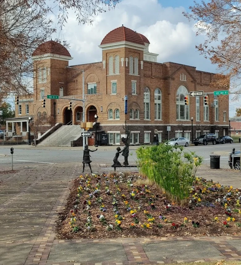 Een historische bakstenen kerk met twee koepeltorens in Alabama, met op de voorgrond een bloemperk en bronzen kinderstandbeelden