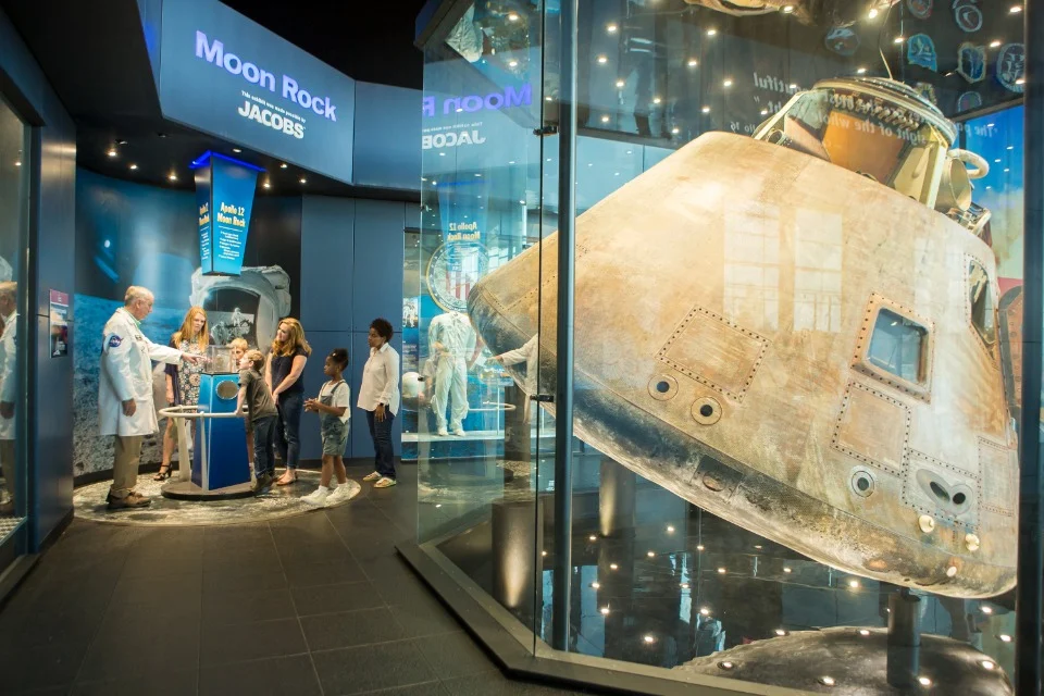Een gids en een groep kinderen bekijken een moonrock naast een grote maancapsule in een ruimtevaartmuseum in Huntsville, Alabama