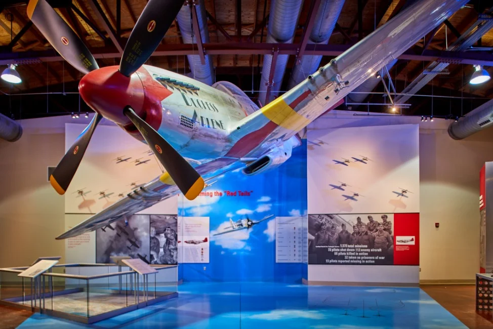 Een gerestaureerd oorlogsvliegtuig met rode neus en propeller hangt boven een expositieruimte in het Tuskegee Airmen museum in Alabama, omringd door informatiepanelen en historische foto's