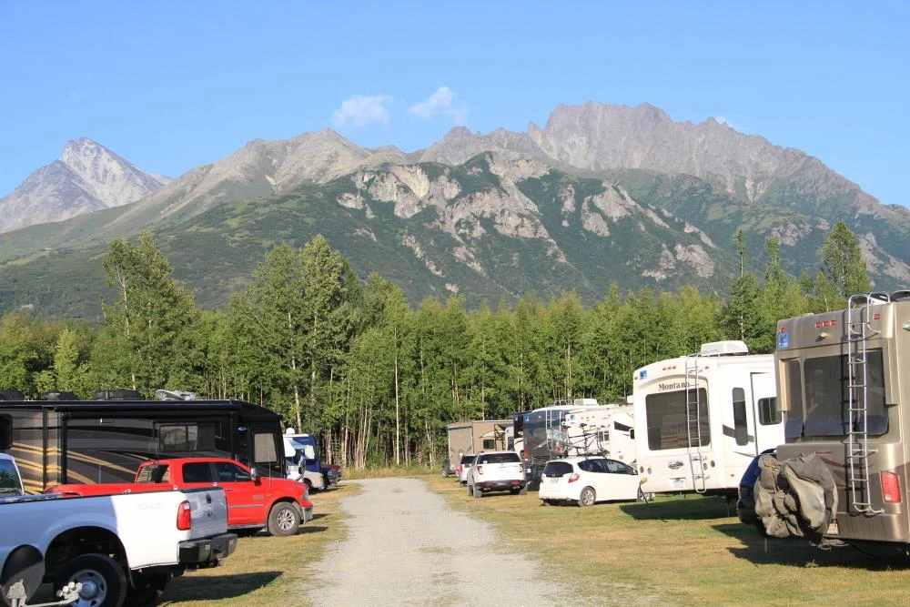 Camperplaats in Alaska met verschillende campers en pickup trucks, omringd door bos en een ruig bergmassief op de achtergrond
