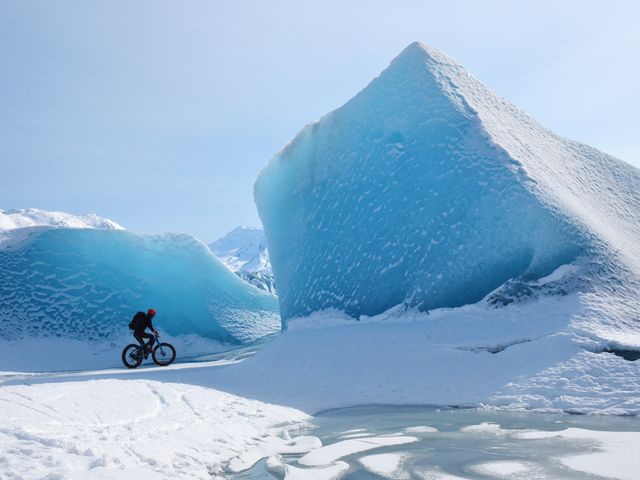 Een gebundelde fietser rijdt op een fatbike over sneeuw langs een grote blauwe ijsberg in Alaska
