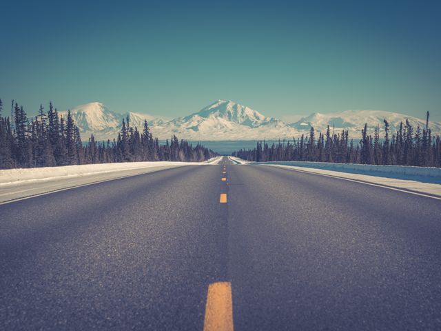 Een verlaten asfaltweg met gele middenstreep loopt door een dennenbos naar besneeuwde bergtoppen in Alaska