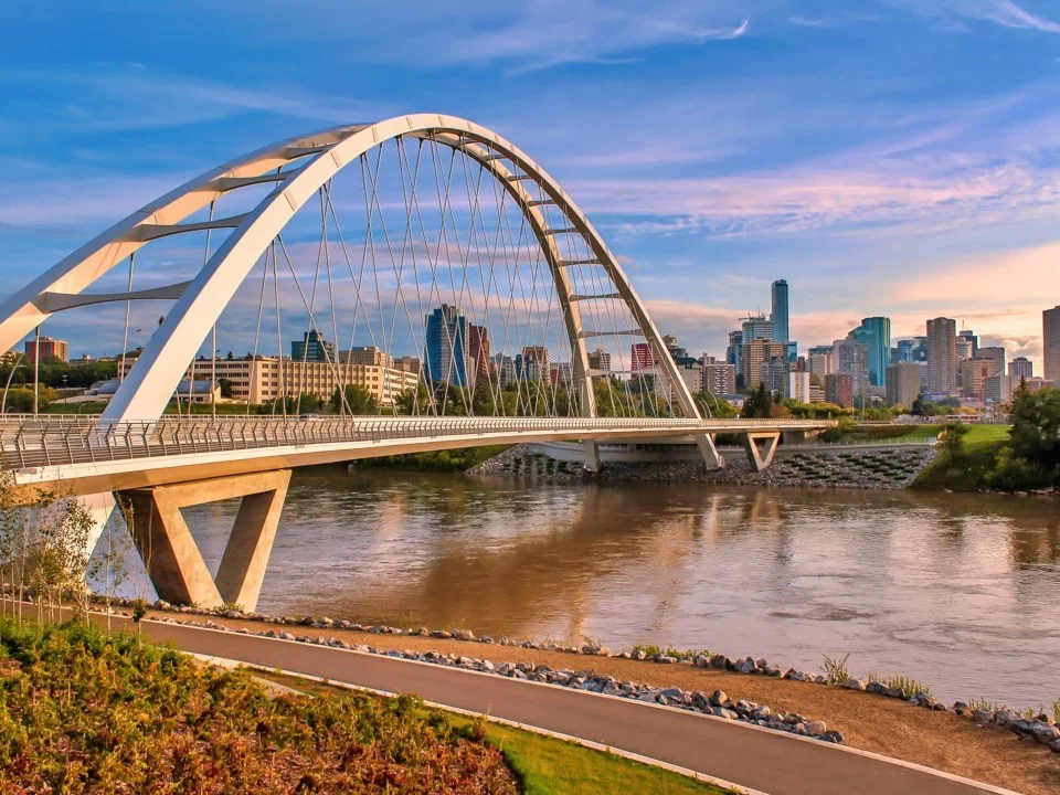 Een moderne witte boogbrug overspant een rivier met bruinig water, met op de achtergrond de skyline van een stad in Alberta bij zonsondergang