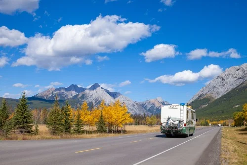 Een witte camper rijdt over een tweebaansweg in Alberta, omringd door herfstbomen en besneeuwde bergtoppen onder een blauwe lucht met wolken
