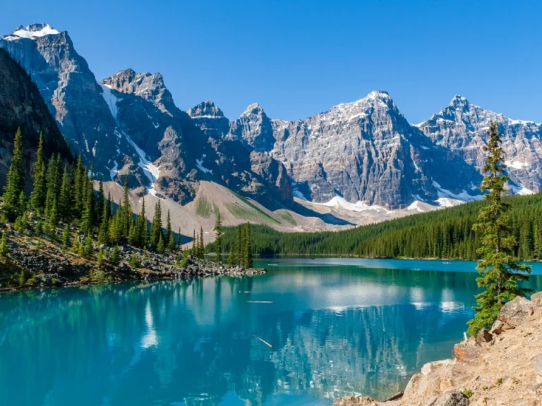 Een helder turkoois meer met dennenbomen aan de oever, omringd door ruige besneeuwde bergtoppen in Alberta