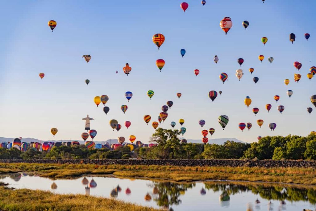 Tientallen kleurrijke heteluchtballonnen stijgen op boven een rivier en groen landschap tijdens de Balloon Fiesta in Albuquerque