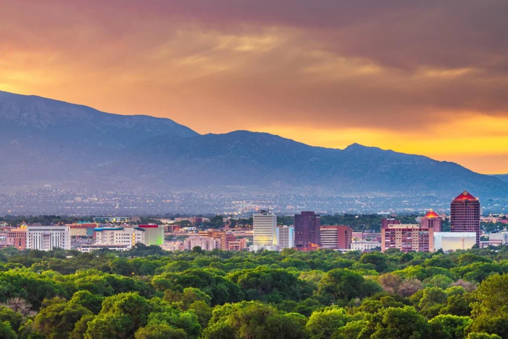 Skyline van Albuquerque met bomen op de voorgrond en de Sandia bergen op de achtergrond onder een oranje avondlucht
