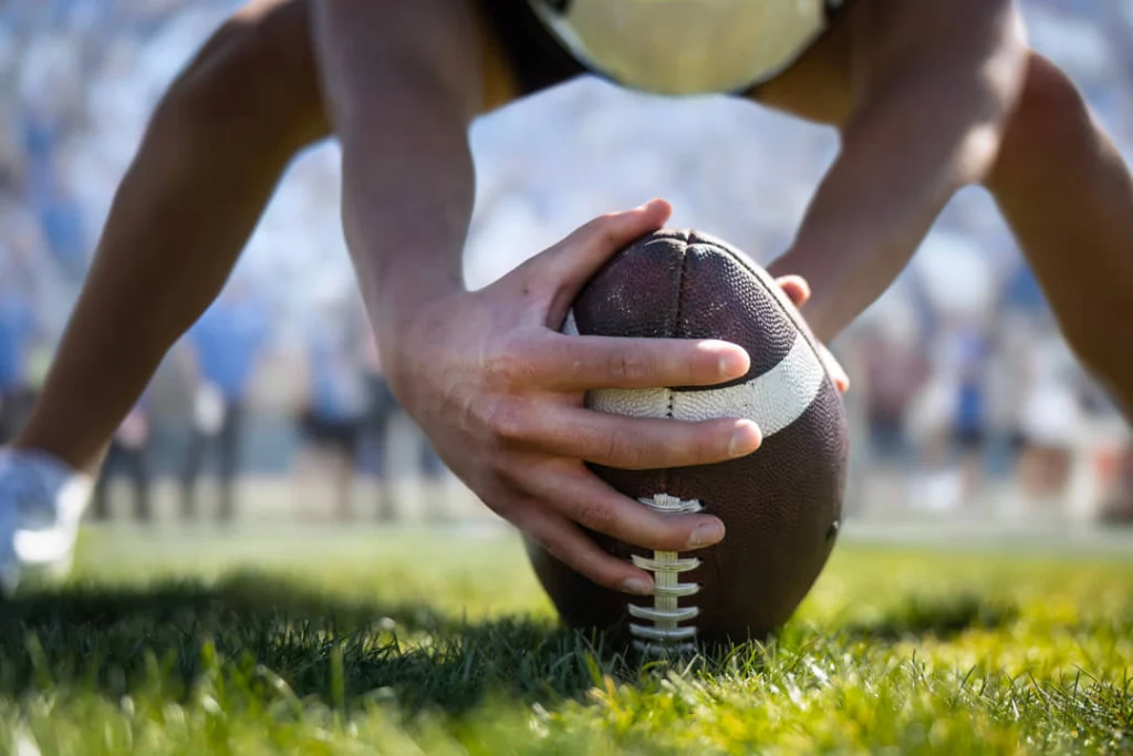 Close-up van de handen van een speler die de American football vastpakt op het gras tijdens de snap