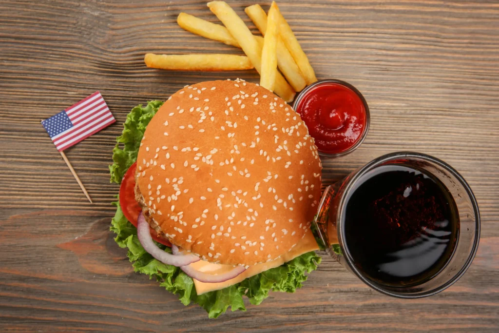 Bovenaanzicht van een hamburger met sesamzaadjesbroodje, sla, tomaat en ui naast friet, ketchup, cola en een klein Amerikaans vlaggetje op een houten tafel